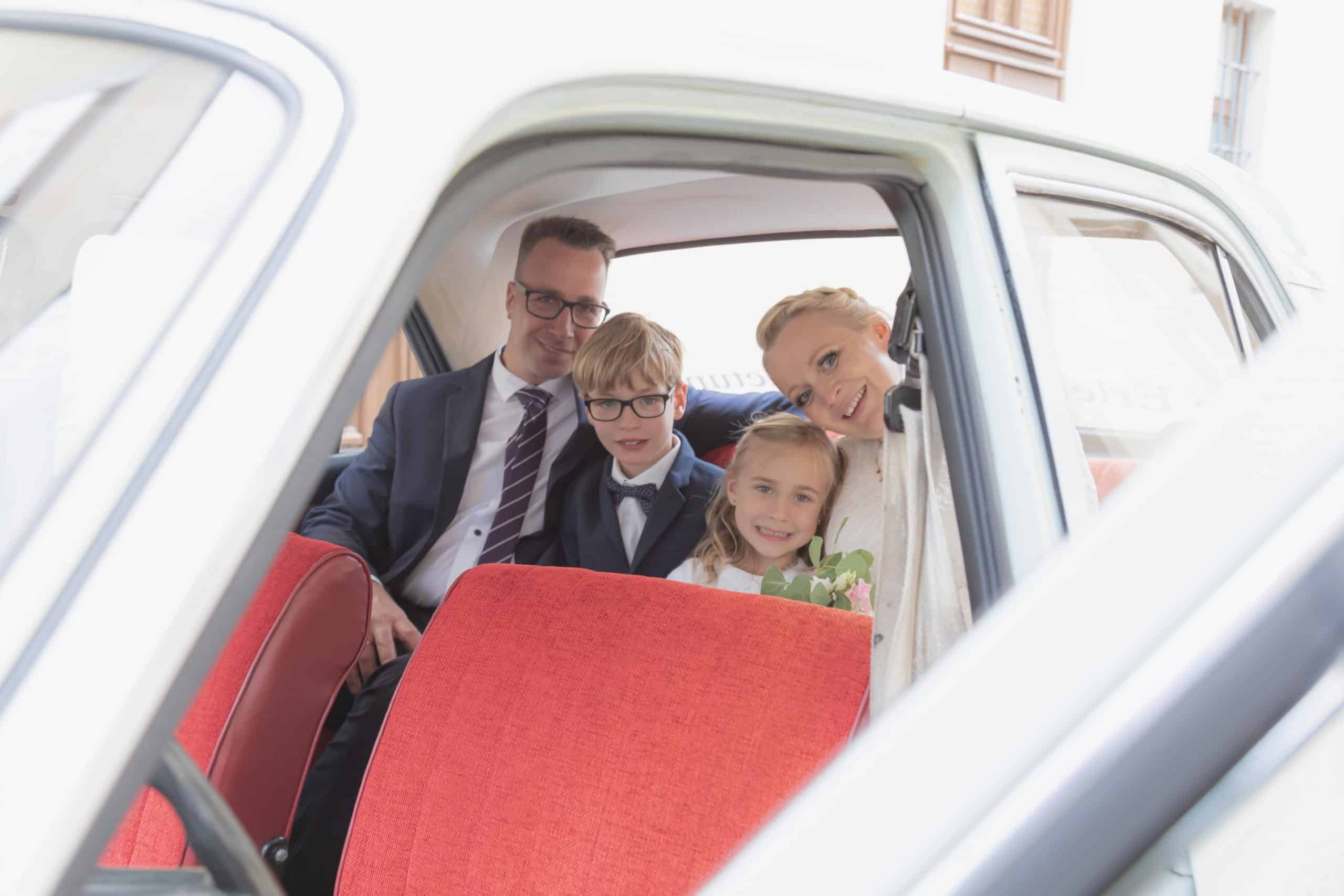 Jan Windisch, Fotograf und Hochzeitsfotograf aus Dresden, Prina, Görlitz, Großenhain und Leipzig, hat hier eine Familie fotografiert. Alle vier Familienmitglieder hocken auf der Rückbank eines Autos und lächeln in die Kamera. Die zwei Erwachsenen, sowie die zwei Kinder sind sehr schick gekleidet.