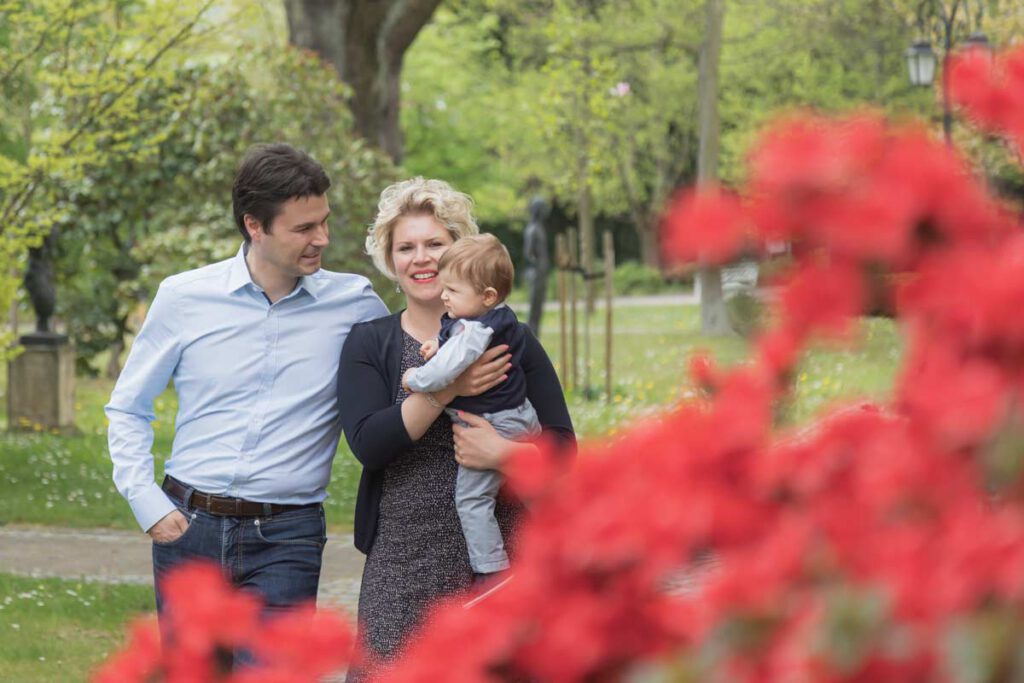 Jan Windisch, Fotograf und Hochzeitsfotograf aus Dresden, Prina, Görlitz, Großenhain und Leipzig, hat hier eine Familie fotografiert. Verschwommen im Vordergrund sieht man einen Busch rote Rosen aus der unteren rechten Ecke ragen. Die Eltern haben laufen auf die Kamera zu und sie trägt ihr Kind im Arm.