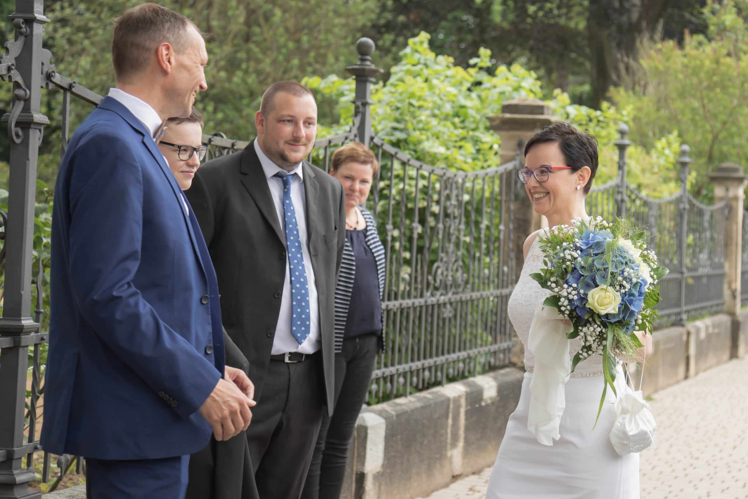 Jan Windisch, Fotograf und Hochzeitsfotograf aus Dresden, Prina, Görlitz, Großenhain und Leipzig, hat hier die Braut und Familie und Freunde fotografiert. Drei Personen reden auf dem Foto mit der Braut. Sie trägt einen Blumenstrauß in der linken Hand.