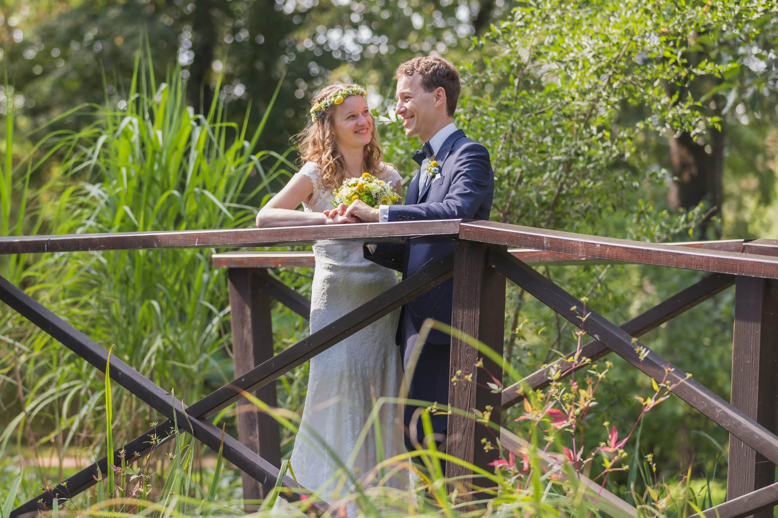 Jan Windisch, Fotograf und Hochzeitsfotograf aus Dresden, Prina, Görlitz, Großenhain und Leipzig, hat hier ein Hochzeitspaar fotografiert. Beide stehen in einem Park auf einer Brücke, mitten im Grünen. Sie schaut ihn an und hat einen Blumenstrauß in der Hand.