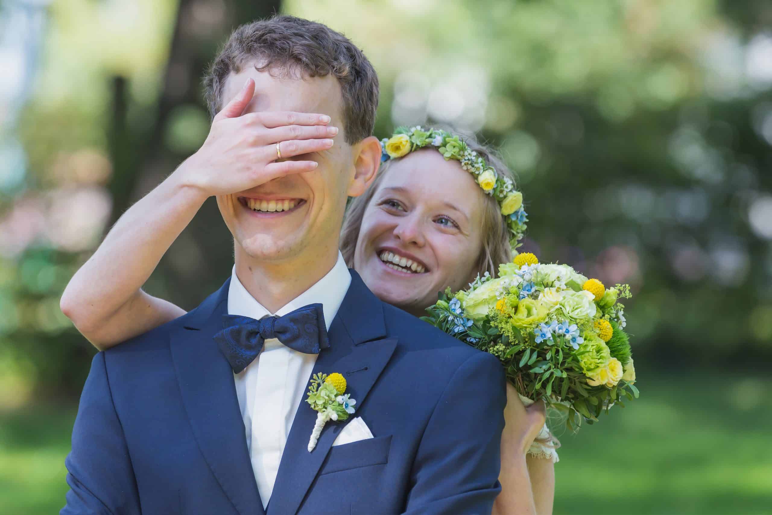 Die Braut hält ihrem Mann die Augen zu und sie lächelt dabei...