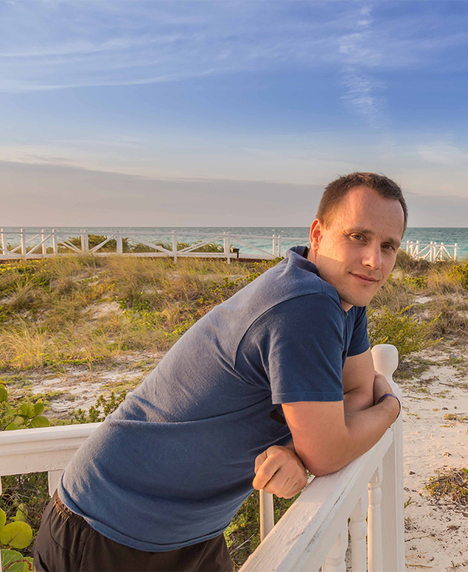 Hochzeitsfotograf Jan Windisch lehnt lässig an einem Holzgeländer und schaut in die Kamera. Im Hintergrund sieht man das Meer von Kuba.