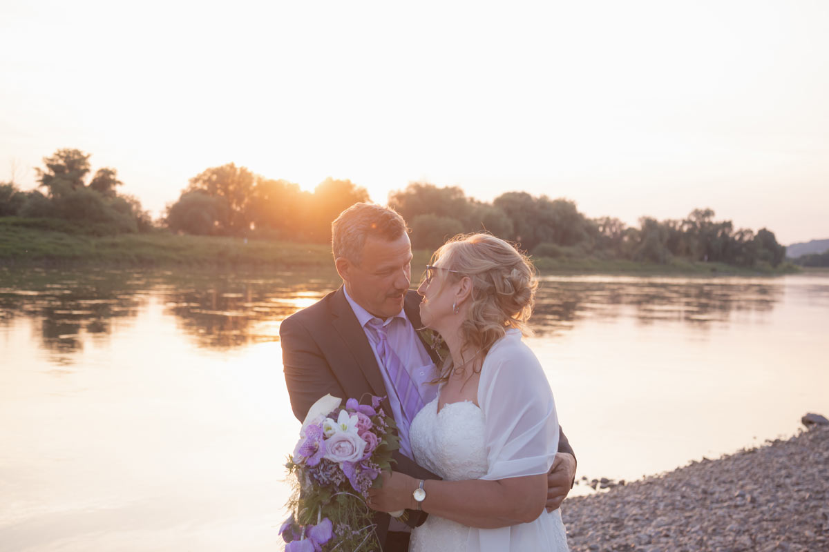 Jan Windisch, Fotograf und Hochzeitsfotograf aus Dresden, Prina, Görlitz, Großenhain und Leipzig, hat hier ein Brautpaar fotografiert. Beides stehen nebeneinander vor einem See, Arm in Arm und sie hält einen Blumenstrauß in der Hand.