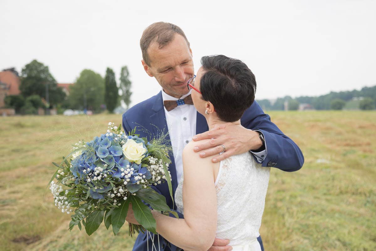 Jan Windisch, Fotograf und Hochzeitsfotograf aus Dresden, Prina, Görlitz, Großenhain und Leipzig, hat hier ein Brautpaar fotografiert. Die Frau wird von hinten dargestellt und hält einen blauen Blumenstrauß in der linken Hand. Der Bräutigam steht hinter der Braut und wird von vorne dargestellt. Das Paar schaut sich verliebt in die Augen