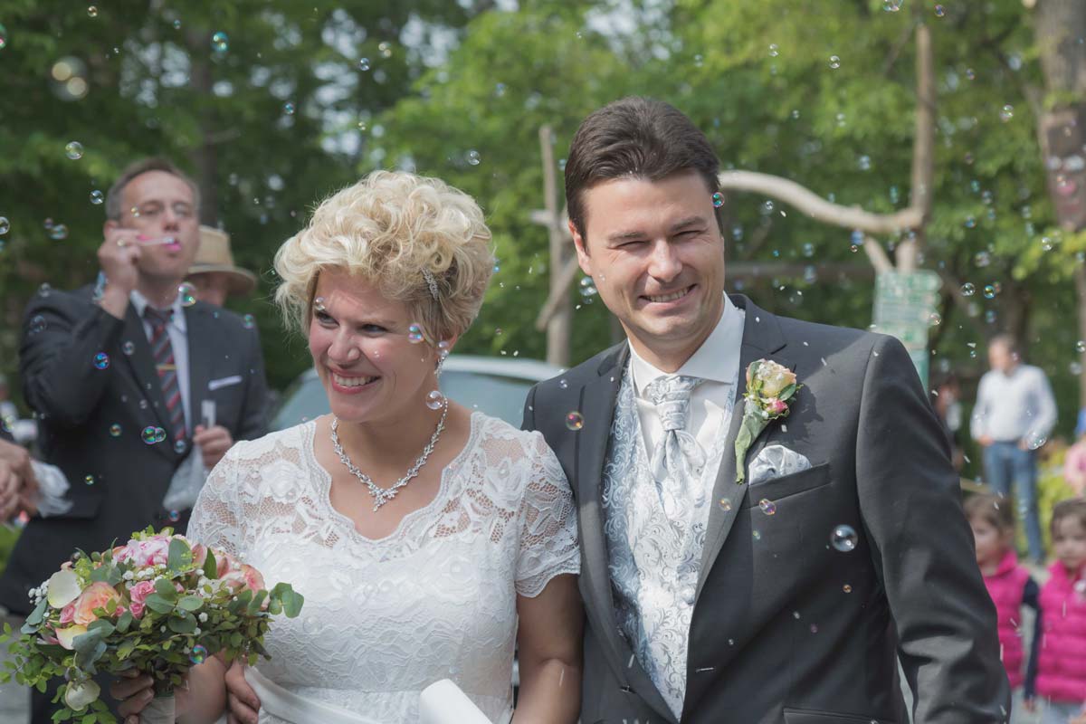 ein glückliches Hochzeitspaar wird von den Gästen empfangen die links und rechts stehen. Überall werden Seifenblasen gemacht und alle freuen sich mit dem Brautpaar und genießen diesen wundervollen Tag