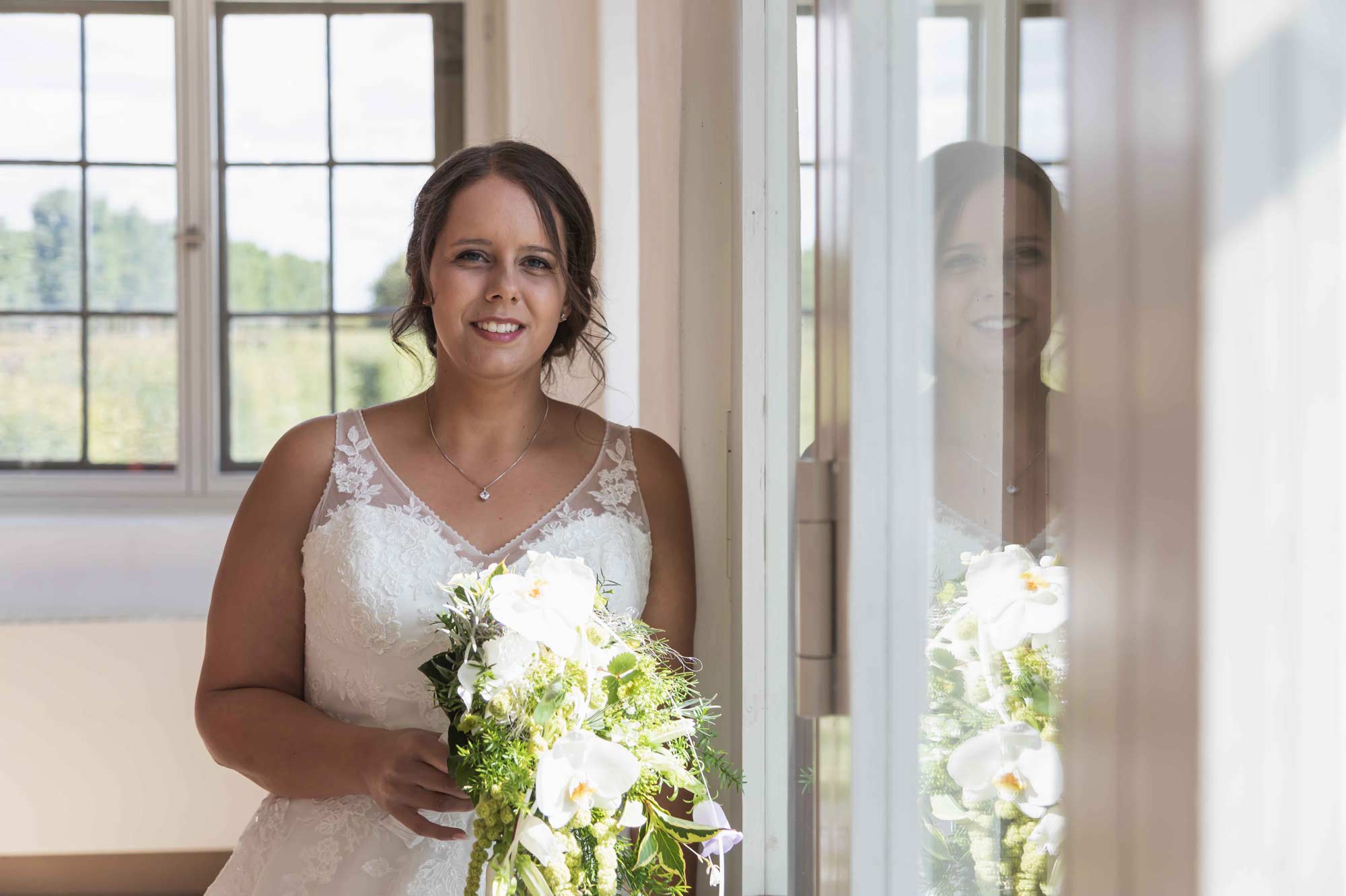 die Braut steht an einem hellen Fensterfront und hält in ihren Händen den Brautstrauß. Sie sieht sehr glücklich aus und wartet auf ihren zukünfitgen mann